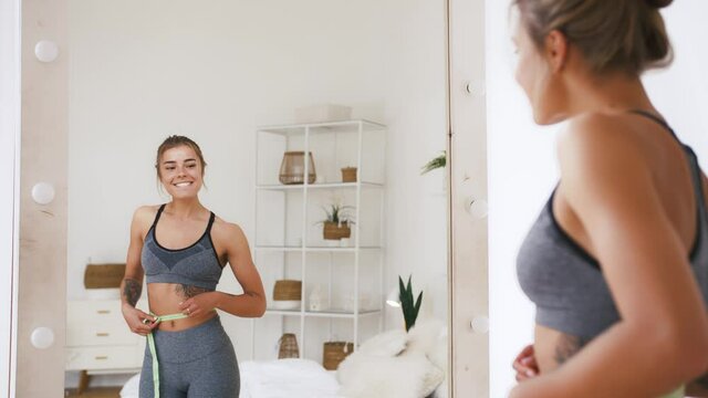Young lady measures waist with measuring tape. Rejoicing by weight loss, looking at mirror in white bedroom. Dieting, eating disorder. Close up