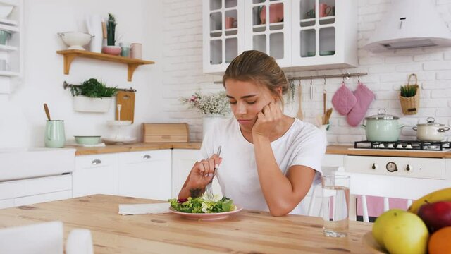 Lady sitting at table, feeling sad and bored with diet not wanting to eat salad. Modern kitchen interior. Healthy nutrition, eating disorder. Close up