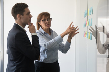 Employees brainstorming, discussing startup ideas, looking on stickers paper on glass wall, confident businesswoman mentor coach training intern, thoughtful businessman pondering strategy