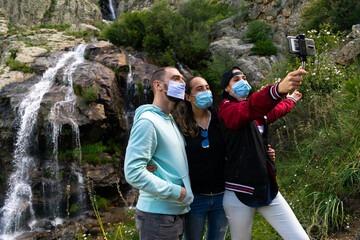 Caucasian people taking a selfie wearing mask in a waterfall. Group of people travelling around Europe during the coronavirus (covid 19) health crisis wearing sanitary mask. Eco turism