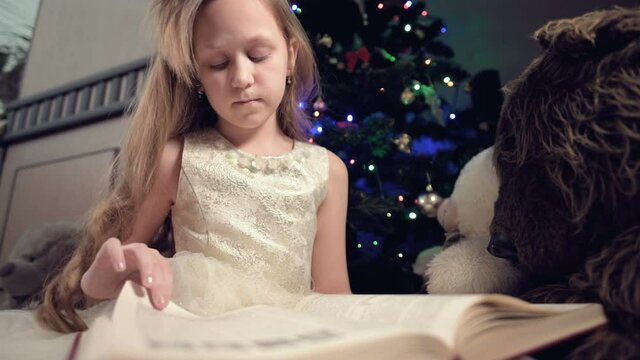 A little blonde girl in a festive dress with a book in her hands sits next to soft toys against the background of a Christmas tree and reads a book leading the page with her place.