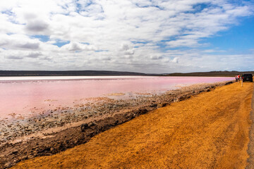 Hutt lagoon: salty lake with pink water due to algae effect. Panoramic view with orange sand and clouds in the sky. People. Location: West Australia WA, Australia.