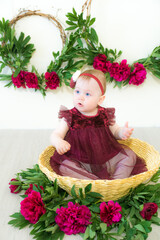 Little cute girl 1 year old in a dress of the color of Marsala sits in a wicker basket with peonies. Spring and flowers. Children's fashion