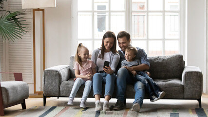 Happy family with children having fun with smartphone, sitting on cozy couch in living room, smiling mother and father with adorable son and daughter looking at phone screen, using mobile apps
