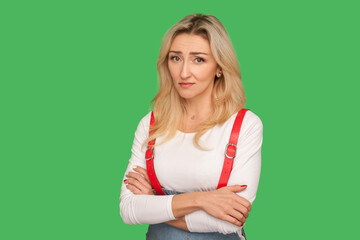 Portrait of unhappy gloomy adult blond woman in stylish overalls standing with crossed hands and looking sadly at camera, feeling helpless desperate. indoor studio shot isolated on green background