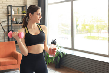 Fototapeta na wymiar Young beautiful woman standing with dumbbell and drink water after exercising. Attractive female bodybuilder working out. Fitness and healthy lifestyle concept.