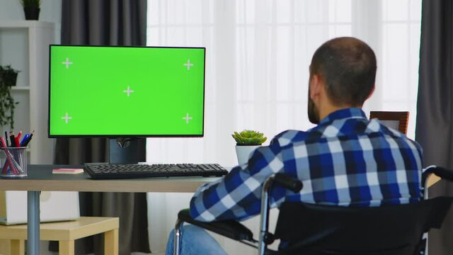 Invalid businessman waving during a video call on computer with green screen. Handicapped invalid paralysed freelancer, immobilized entrepreneur working from home, ilness and disability