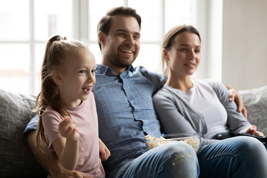 Happy parents with little daughter watching tv, comedy movie or soccer match together, sitting on cozy couch in living room, enjoying leisure time at home, family eating popcorn snacks
