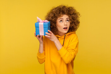 Portrait of funny curious curly-haired woman in urban style hoodie holding present box near ear and...