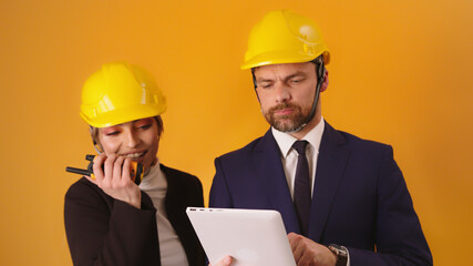 Man and woman professional architects with hard hats, tablet and walkie talkie isolated on orange background.