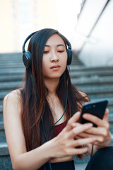 Asian girl in the park listens to music on headphones. Girl during a walk with the phone. Summer time. Chat on the phone.