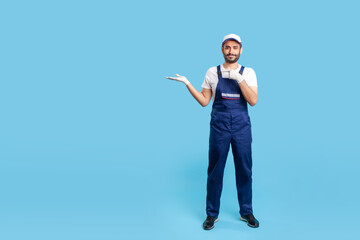 Naklejka premium Full length cheerful worker in blue overalls, cap and protective gloves standing, pointing at empty space on his hand. Profession of service industry, courier or labourer. studio shot isolated