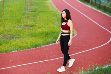 Fototapeta premium Asian girl go in for sports on a sports track on the street. Red treadmill. Girl with headphones. Girl in a sports top and leggings.