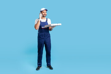 Full length positive young courier in workwear talking on cellphone, holding pizza box and communicating with client. Delivery man answering phone call, accepting order. indoor studio shot isolated