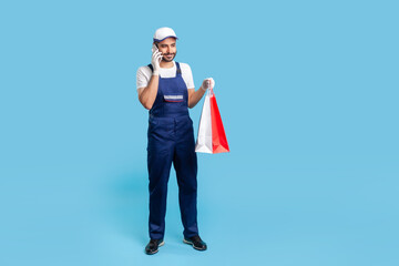 Full length portrait of smiling courier in workwear talking on cellphone, holding shopping bags and communicating with client. Delivery man answering phone call, accepting order at fashion store