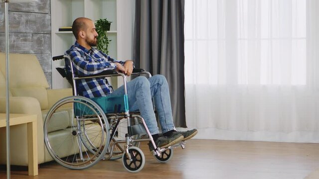 Unhappy man in wheelchair in living room looking at window. Handicapped invalid paralysed person, alone at home, depression and health problems, pensive, sad and unhappy man