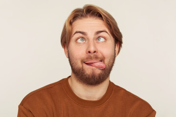 Closeup portrait of dumb fool bearded man looking cross-eyed and showing tongue out, fooling around, making silly face, brainless comical expression. indoor studio shot isolated on gray background