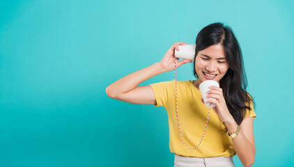 Portrait happy Asian beautiful young woman smile white teeth standing wear yellow t-shirt, She holding paper can telephone for talking, studio shot on blue background with copy space for text