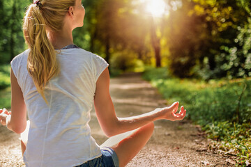 Young blonde woman sitting in lotus position in park and meditating, sunset backround.