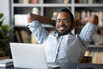 African relaxed guy sit at desk leans on office chair put hands behind head takes break at...