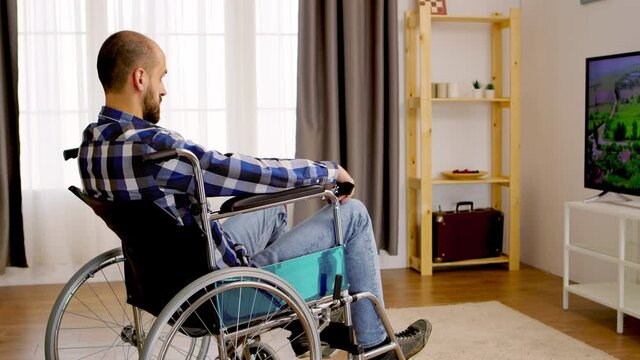 Thoughtful paralyzed man in wheelchair watching tv. Handicapped invalid paralysed person, alone at home, depression and health problems, pensive, sad and unhappy man