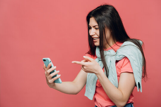 Delighted Young Brunette Dressed In A Pink T-shirt And   Turquoise Sweater Tied Up Around Her Neck, Holds Her Smartphone And Point At It Over Pink Background, With Surprised Face After Video Call