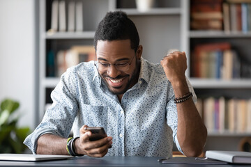 Excited African man sitting at workplace desk looking at smartphone screen read unbelievable...