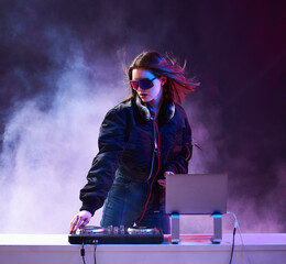 Stylish girl DJ in glasses headphones and a bomber mixes music dancing at a party in the neon light...