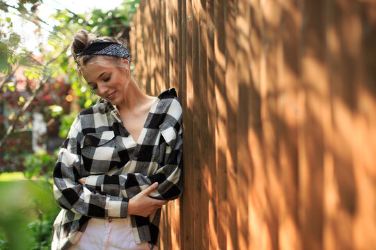 Portrait of a pretty girl in a plaid shirt and bandana standing against a wooden fence in the backyard on a Sunny day.