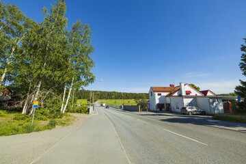 Beautiful nature landscape view. White houses on one side of asphalt road and green birch trees on another side on blue sky background.
