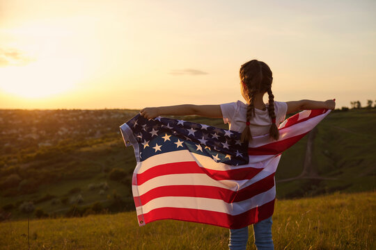 Independence Day USA. Rear view of a kid girl with the flag of America on nature at sunset.