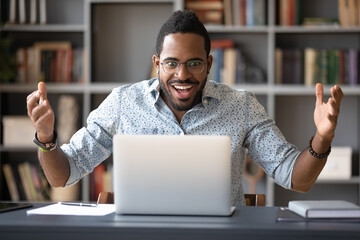 Surprised African guy sitting at desk looking at computer screen makes big eyes feels shocked by unbelievable news, got opportunity new job, low price discounts sale, lottery or betting win concept - obrazy, fototapety, plakaty