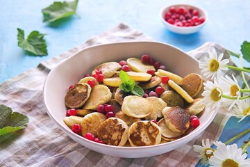 Mini pancakes with red currants in a beige bowl on a blue concrete background.