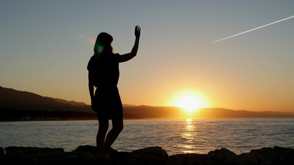 Silhouette of a young woman stands on Marbella seashore at sunrise. Female waving a hand welcomes...