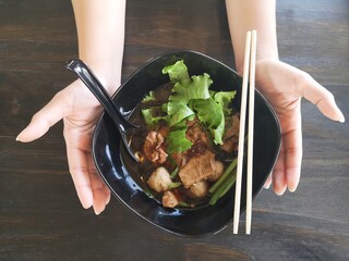 Two hands hold the black bowl of Rice noodles soup with beef, meat ball and lettuce on topping, wood background, thai noodles style 