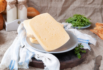 A large piece of semi-hard cheese on a gray plate. On a brown wooden board and a linen towel. In the background are bottles of milk, bread. Arugula and croutons. Background of gray linen. Close-up.