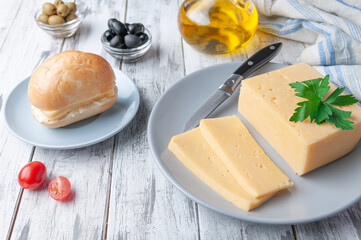 A piece of semi-hard cheese is cut into slices. Lies on a gray plate. Garnished with parsley. In the background a bun with cheese and butter, olives and olive oil. Gray wooden background. Close-up.
