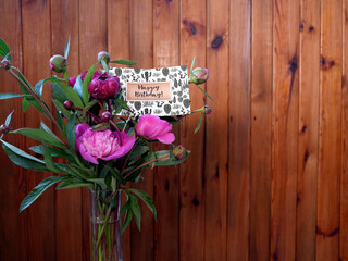 Pink flowers peonies in a vase on a wooden background with a happy birthday card. The photo was taken with natural daylight.