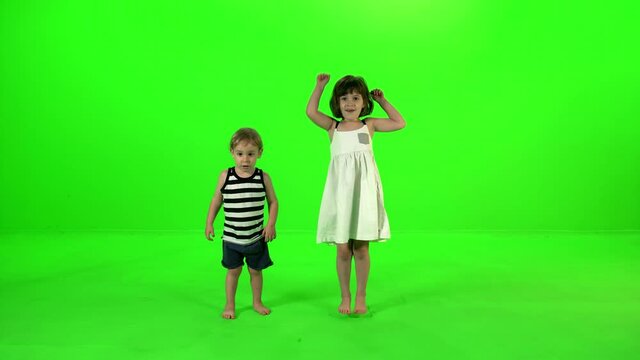 A Cute little girl and a toddler boy dance, cheer and jump, boy leaves the girl to jump alone, over green screen.