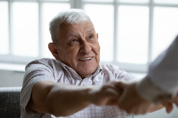 Fototapeta na wymiar Close up smiling older man holding doctor hands, having pleasant conversation, receiving psychological help and care, female caregiver therapist gp supporting mature senior patient at meeting