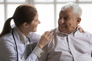 Close up caring female doctor wearing uniform with stethoscope talking to smiling older man, young woman caregiver supporting elderly patient, touching shoulders, warm relationship, healthcare
