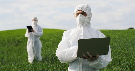 Male Caucasian farmer biologist in white protective costume and goggles standing in green field and...