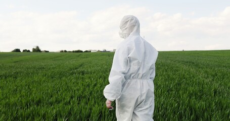 Rear of Caucasian male farmer ecologist in white protective costumes and goggles walking in green...