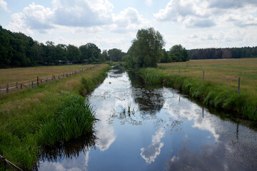 The river Aller flows sluggishly through the meadows near Gifhorn, with fences at the edge and individual bushes and trees, cloud reflections in the water