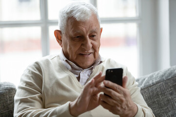 Close up smiling older man using phone, looking at screen, sitting on cozy couch at home, happy mature male holding smartphone, browsing mobile device apps, writing message on cellphone