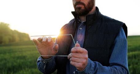 Close up of glass futuistic gadget in hands of male fermer who standing in green field in summer. Hi-tech technology of transparent device. Man tapping on tablet of future. Augmented reality.