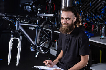 Fototapeta na wymiar Stylish man with beard, cycle mechanic takes notes, checklist before repairing and servicing bike in bicycle shop workshop. Caucasian male in bicycle shop writing on clipboard. Assessing bike supply