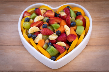 Close-up of a mixed colorful fruit salad in a white ceramic plate heart shaped. Wooden background