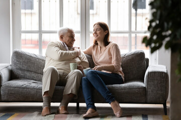 Happy older man and adult daughter drinking tea or coffee together, sitting on cozy couch, smiling young woman and mature father having fun, enjoying leisure time, talking, sharing news