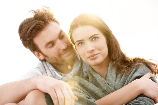 Portrait of young love couple sitting outdoor in sun light, sharing a blanket, smiling. Summer and lens flare effect. 